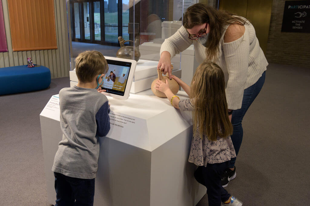 Museum visitors interact with a small touch kiosk an model of an ancient peruvian ceramic artifact