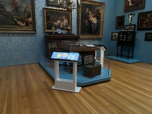 A digital touch kiosk stands in front of a collection of 17th century European furniture