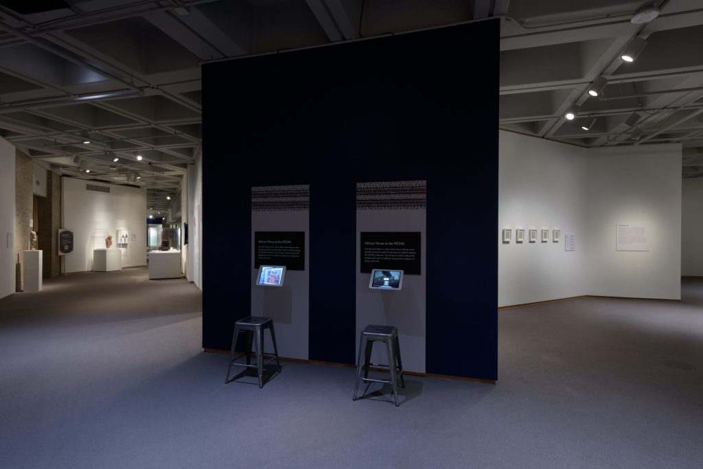A pair of small touchscreen kiosks displaying video content are pictured in an empty art museum gallery